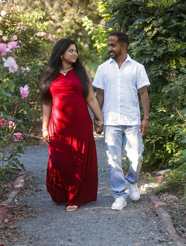 Expecting couple walking hand in hand along a rose garden path at golden hour