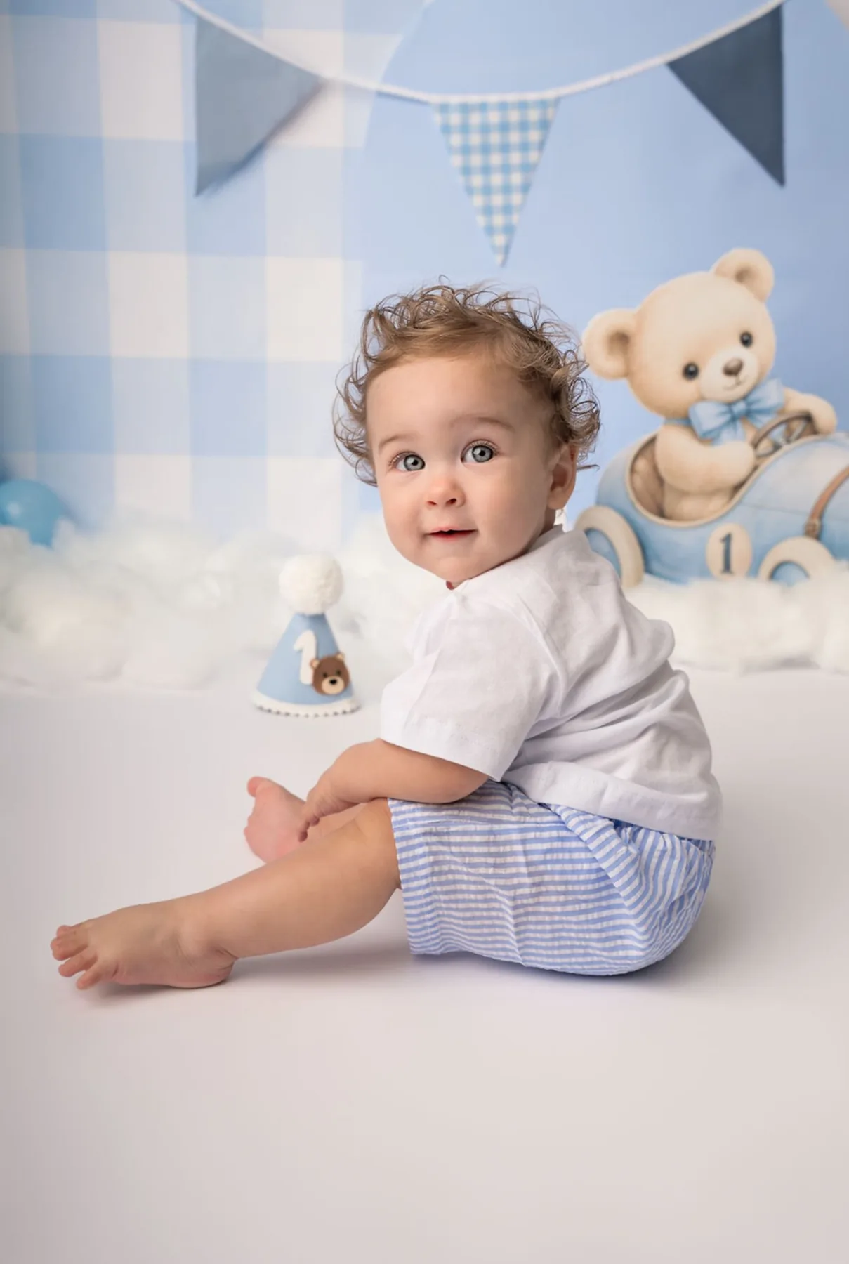 One year old boy in blue gingham shorts in front of a teddy bear first birthday backdrop