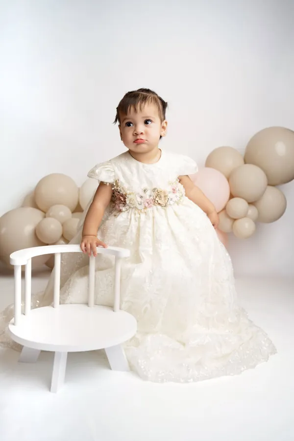 Little girl in a white lace gown standing beside a small white chair and neutral balloons