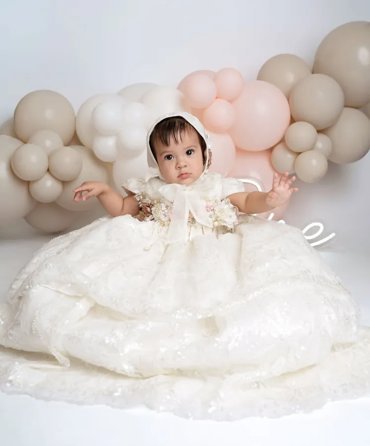 Baby girl in a lace bonnet and flowing white gown among blush and cream balloons