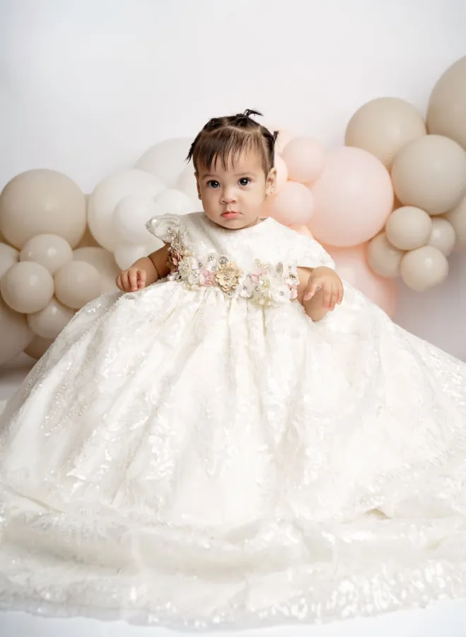 Baby girl in a white gown with a floral waistband in front of blush and cream balloons
